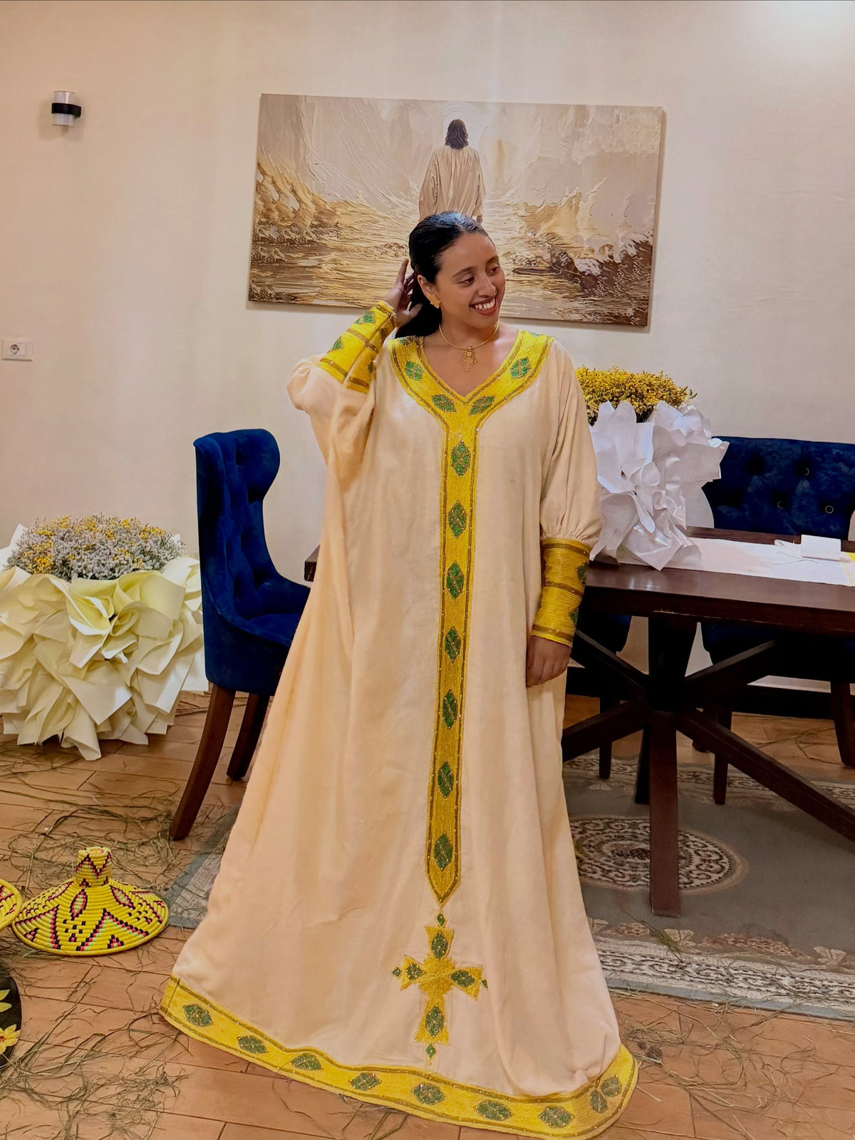 A woman wearing a long cream-colored traditional traditional clothing  with yellow and green embroidery, standing in an indoor room with a table, chairs, and wall art in the background.