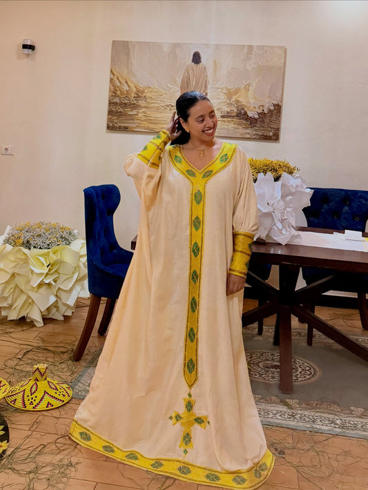 A woman wearing a long cream-colored traditional traditional clothing  with yellow and green embroidery, standing in an indoor room with a table, chairs, and wall art in the background.