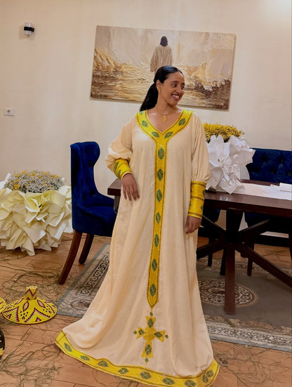 A woman wearing a long cream-colored traditional clothing  with yellow and green embroidery, standing in an indoor room with a table, chairs, and wall art in the background.