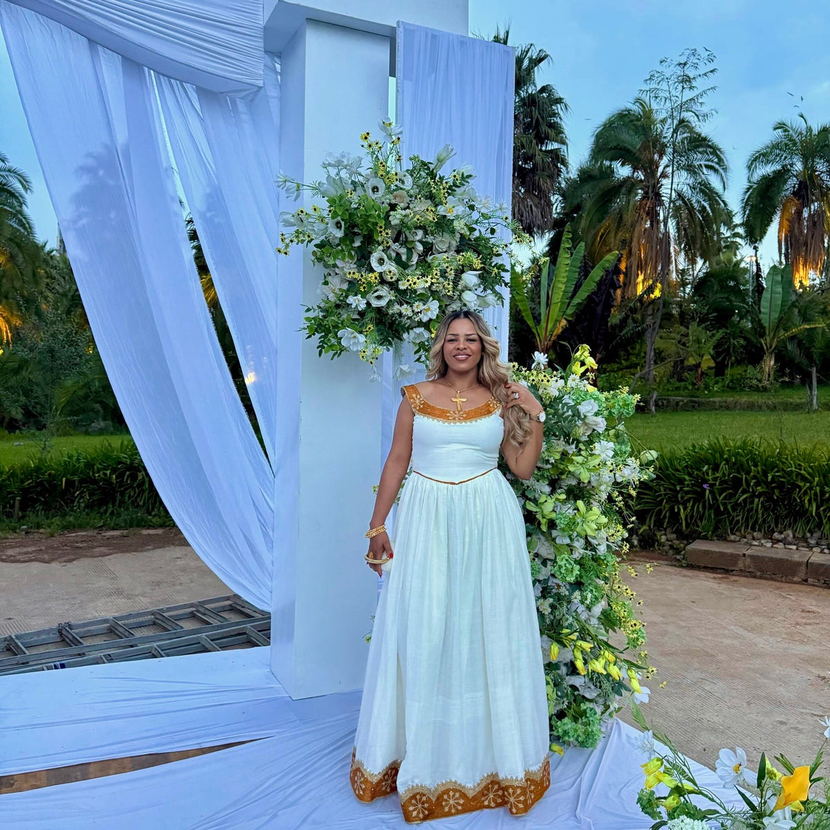 A woman stands outdoors in front of a decorated white arch with draped fabric and floral arrangements, wearing a sleeveless white Ethiopian national dresses with gold trim and a gold embroidered hem.