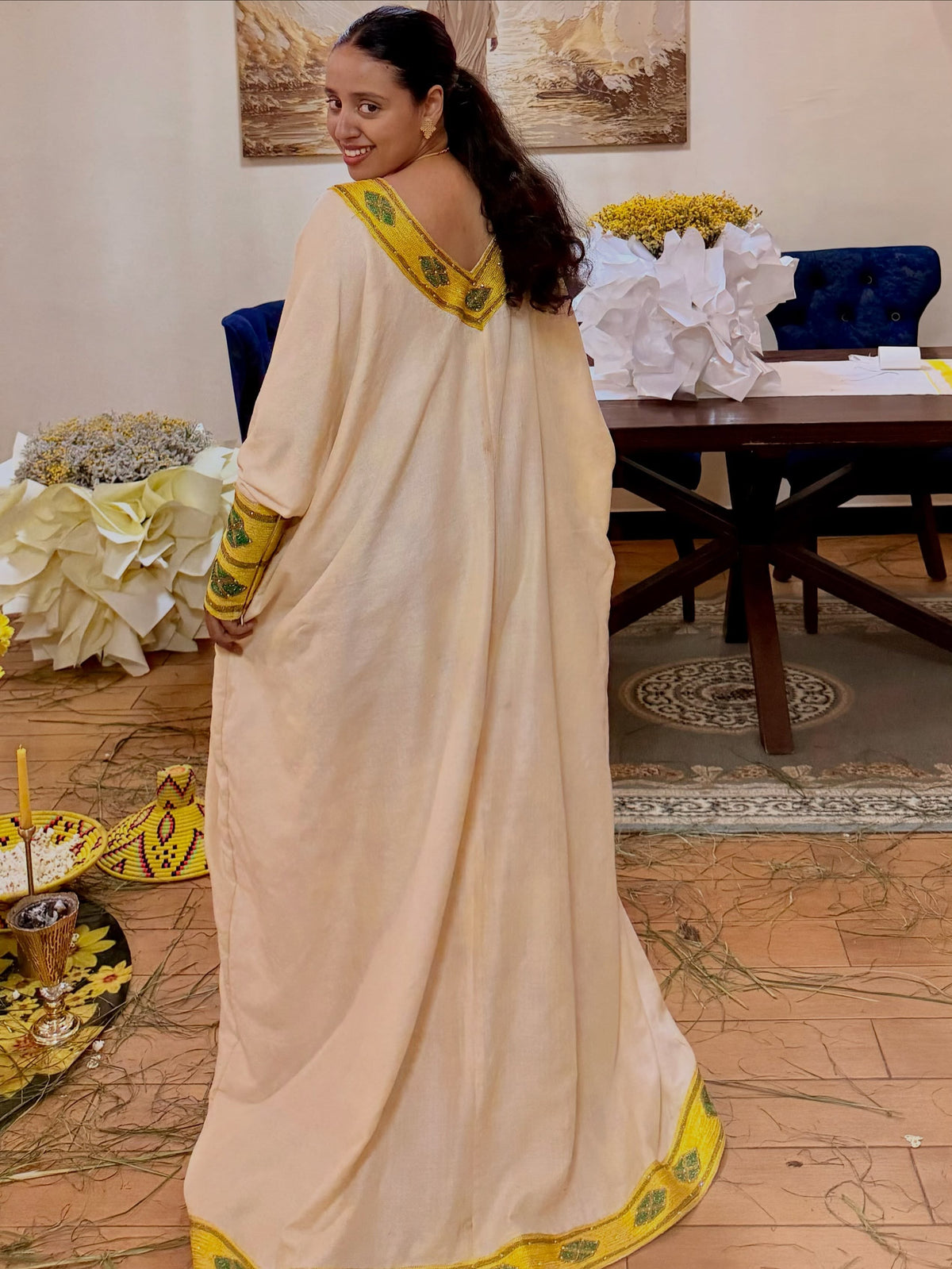 A woman wearing a cream-colored traditional  clothing  with yellow and green border details, photographed in an indoor setting with a table and decorative flowers in the background.