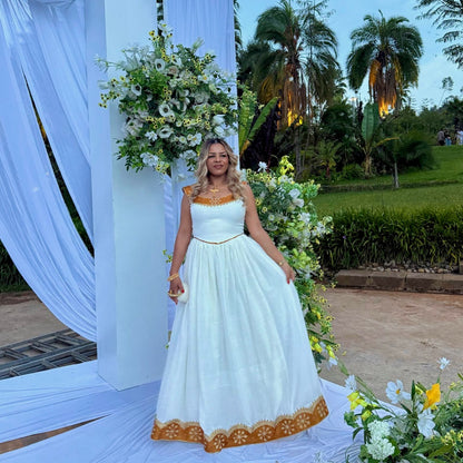 A woman stands outdoors in a white sleeveless maxi Ethiopian national dresses with gold trim and a gold embroidered hem, posed in front of a white draped event arch with floral arrangements and palm trees in the background.