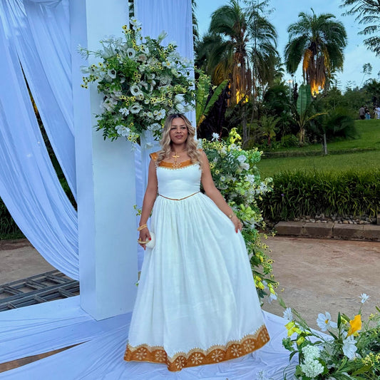 A woman stands outdoors in a white sleeveless maxi Ethiopian national dresses with gold trim and a gold embroidered hem, posed in front of a white draped event arch with floral arrangements and palm trees in the background.
