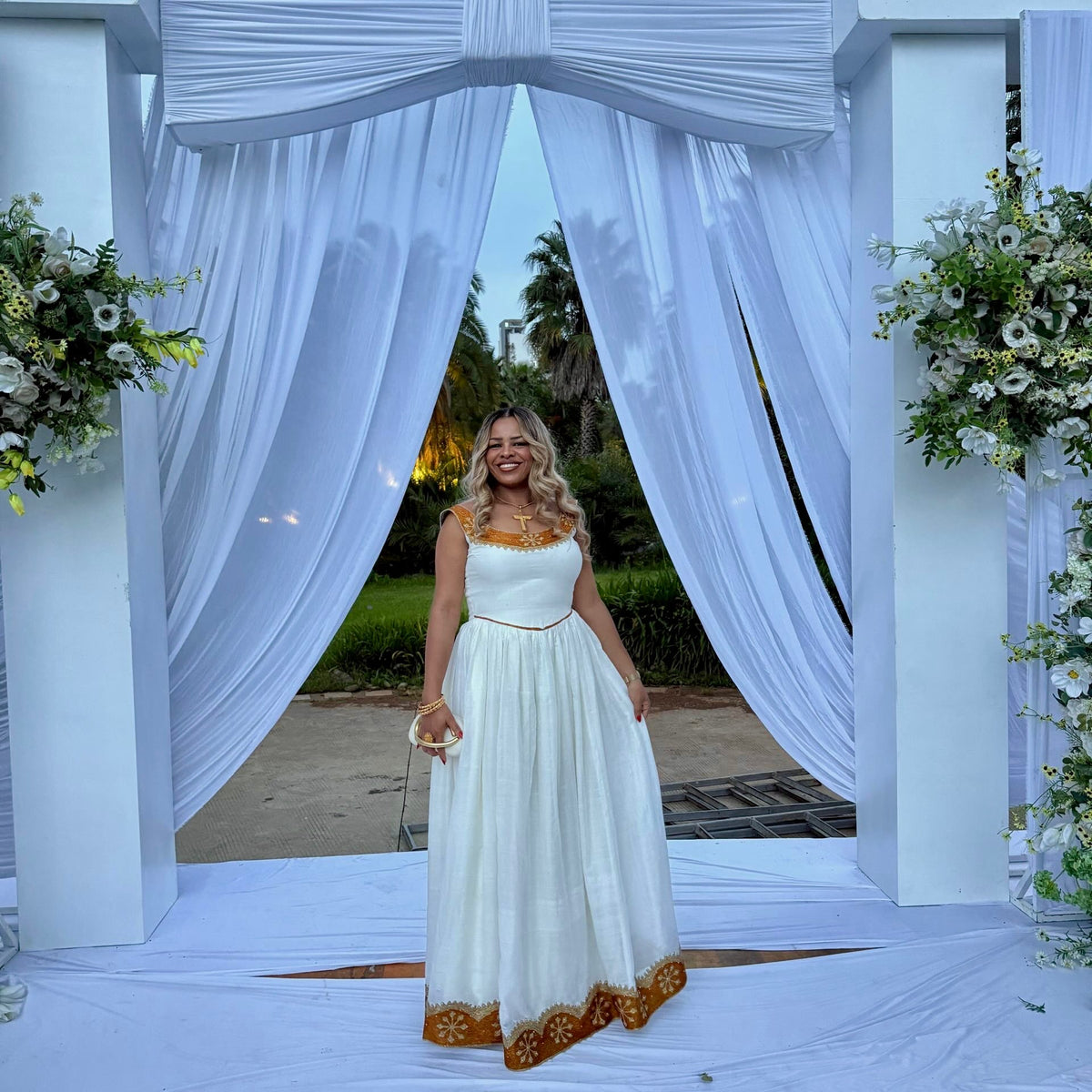 A woman wearing a white sleeveless maxi Ethiopian national dresses with gold embroidery and a gold waistband stands under a white draped wedding arch with floral arrangements.