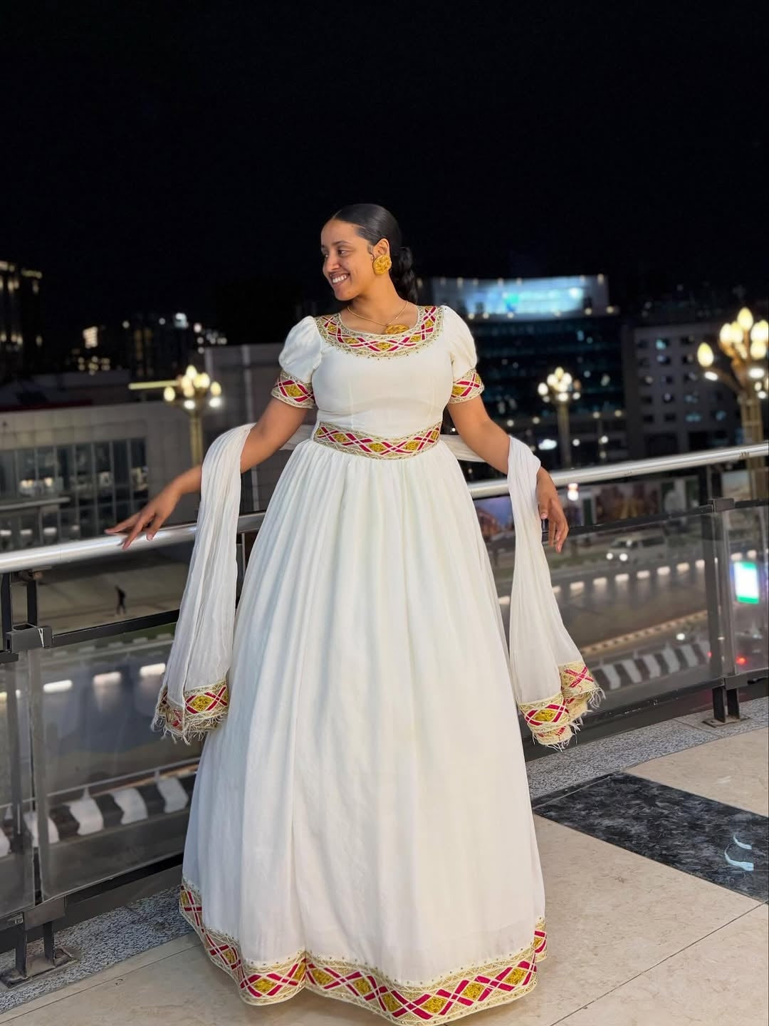 Woman in a white traditional Simple Gold Habesha Libs with colorful patterns standing on a rooftop at night.