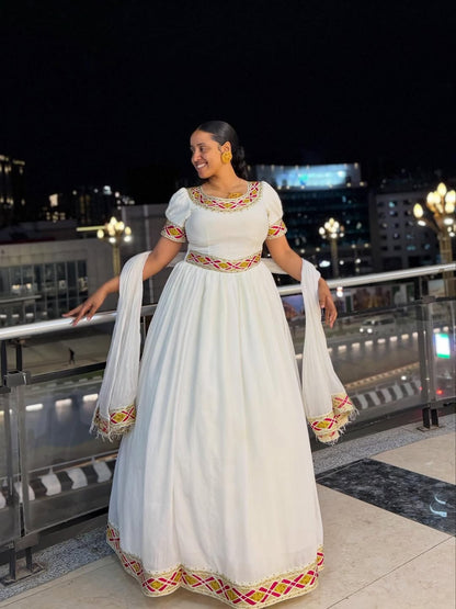 Woman in a white traditional Simple Gold Habesha Libs with colorful patterns standing on a rooftop at night.