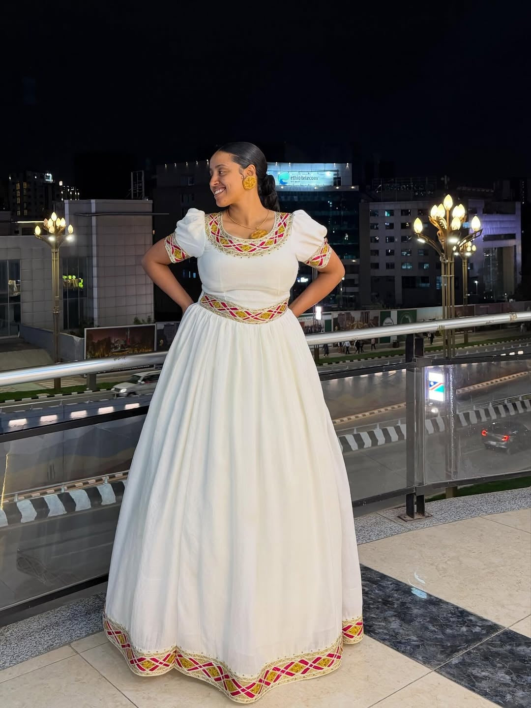 Woman in a white traditional Simple Gold Habesha Libs with colorful patterns standing on a rooftop at night.