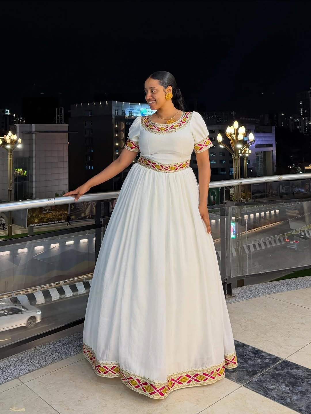 Woman in a white traditional Simple Gold Habesha Libs with colorful patterns standing on a rooftop at night.