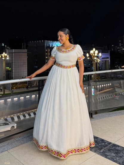 Woman in a white traditional Simple Gold Habesha Libs with colorful patterns standing on a rooftop at night.