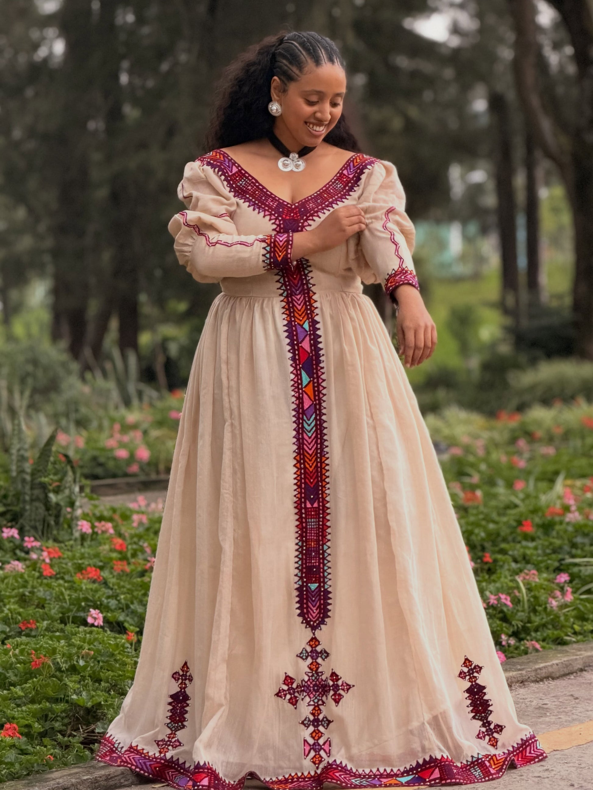 A woman wearing a beige maxi  Ethiopian Kemis  with colorful red, purple, and blue geometric embroidery, standing in a garden setting.