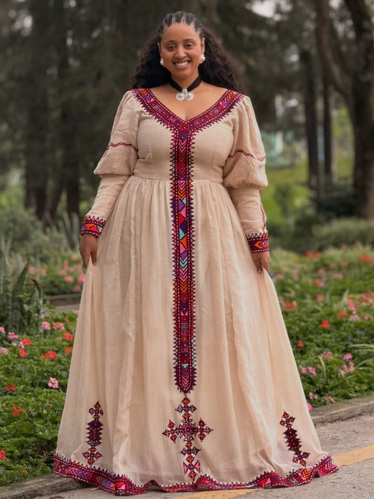 A woman wearing a beige long-sleeve maxi  Ethiopian Kemis with colorful red, purple, and blue geometric embroidery, standing in a garden setting.