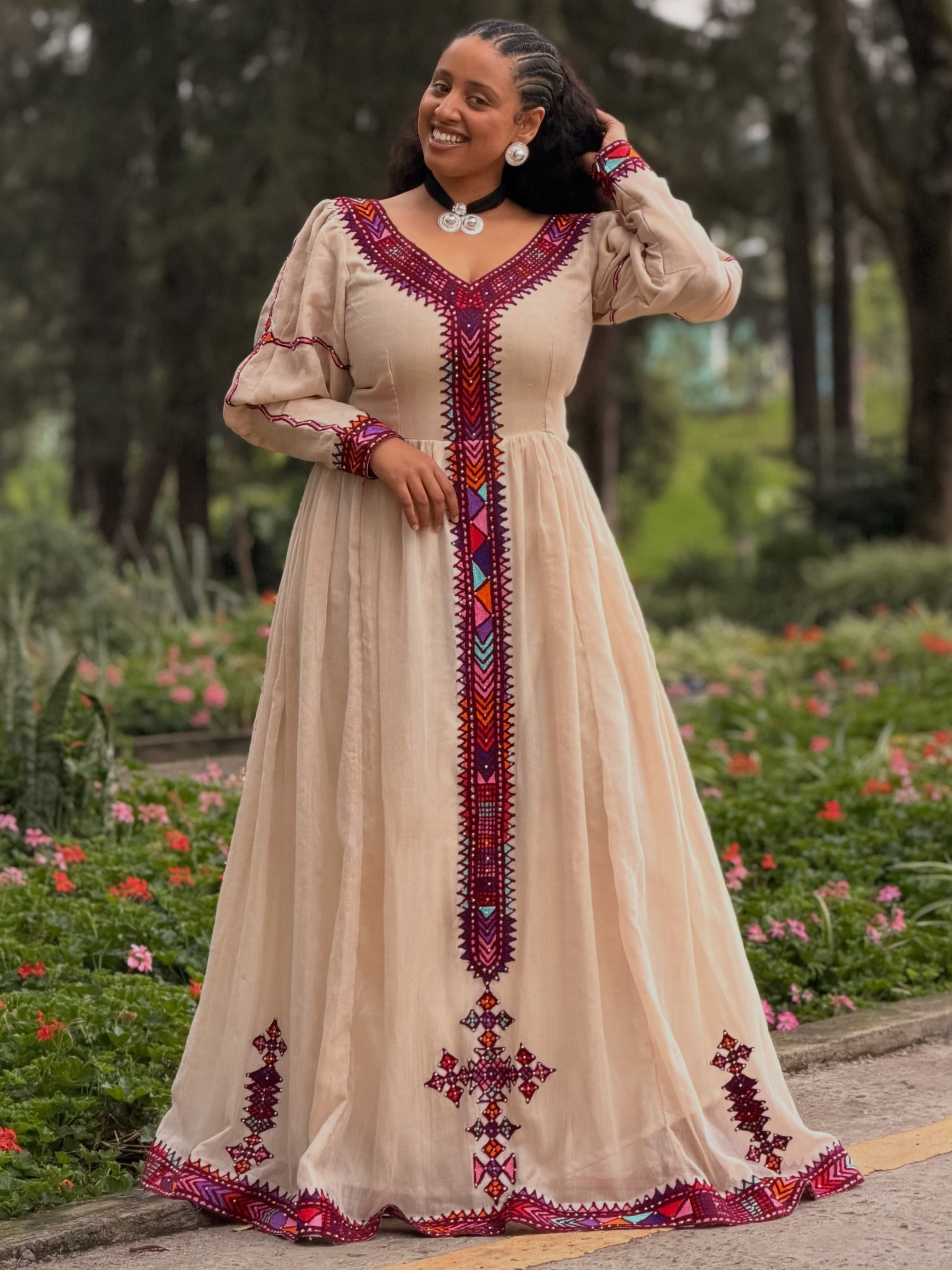 A woman wearing a beige  Ethiopian Kemis  with red, purple, and blue geometric embroidery, standing in a garden setting.