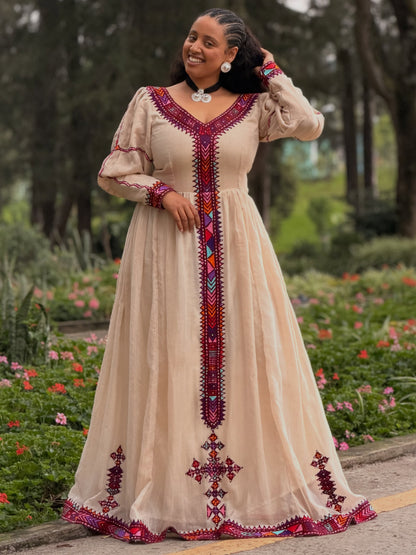 A woman wearing a beige  Ethiopian Kemis  with red, purple, and blue geometric embroidery, standing in a garden setting.