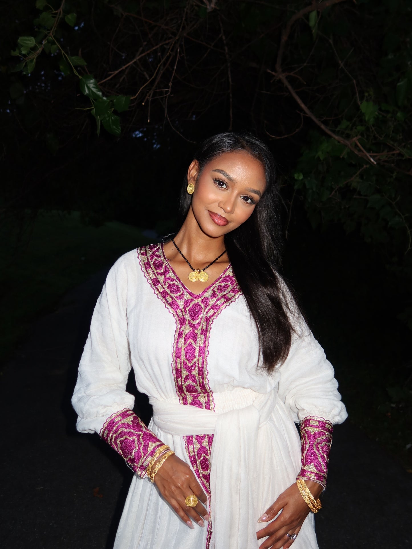 A woman wearing a white long-sleeve ethiopian dress with magenta and gold embroidered patterns, standing outdoors at night against a dark background.