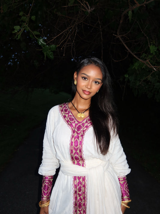 A woman wearing a white long-sleeve ethiopian dress with magenta and gold embroidered patterns, standing outdoors at night against a dark background of trees.