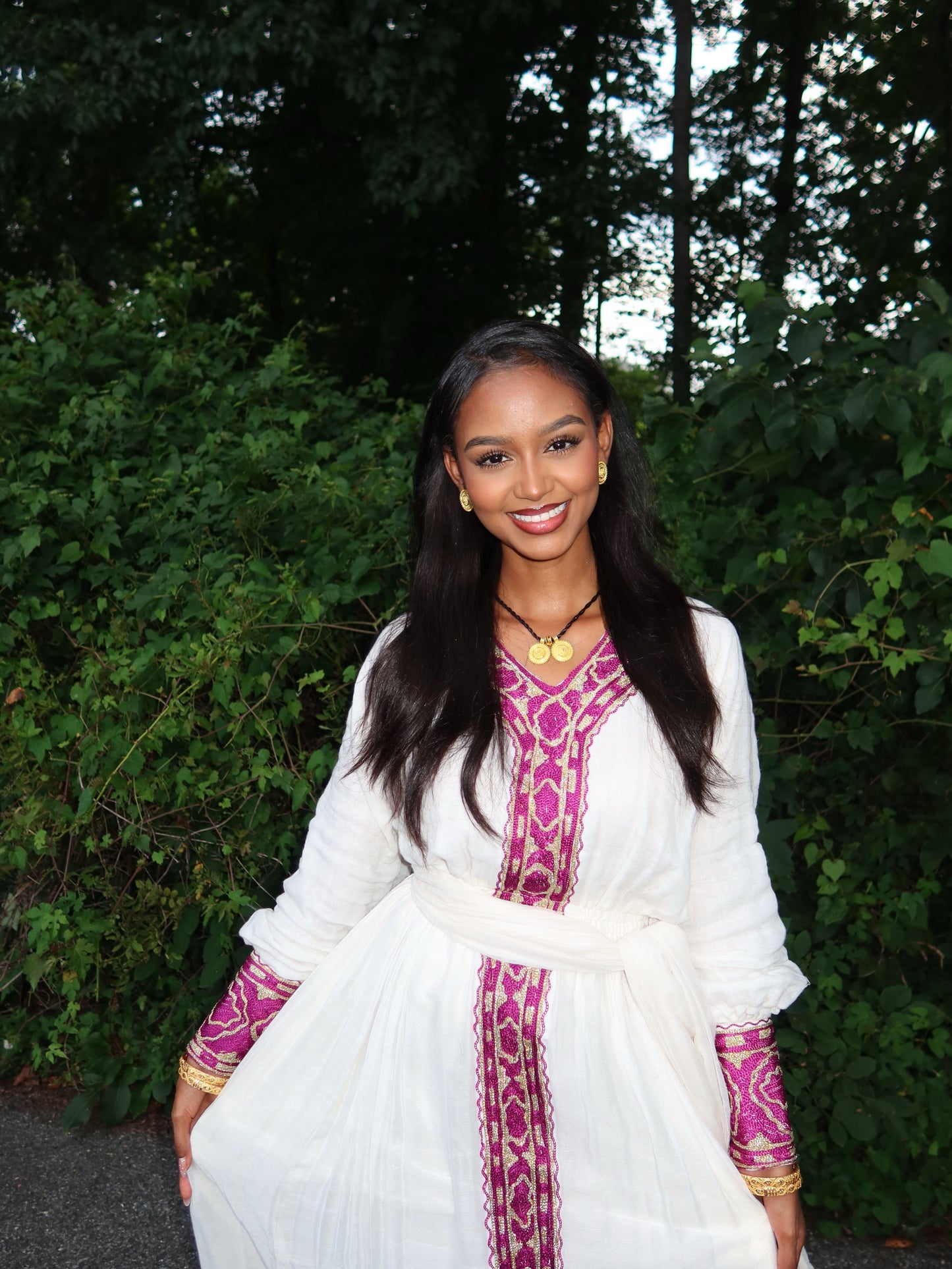 A woman wearing a white long-sleeve ethiopian dress with magenta and gold embroidered patterns, standing outdoors in front of a green leafy background.