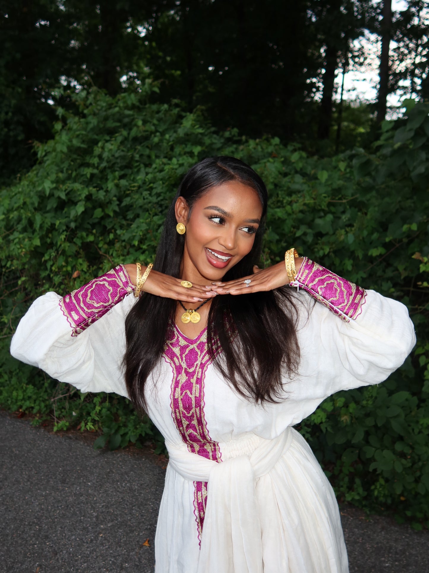 A woman posing outdoors in a white long-sleeved ethiopian dress with magenta and gold embroidered details on the neckline and cuffs, accessorized with gold jewelry.