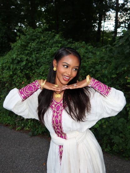 A woman posing outdoors in a white long-sleeved ethiopian dress with magenta and gold embroidered details on the neckline and cuffs, accessorized with gold jewelry.