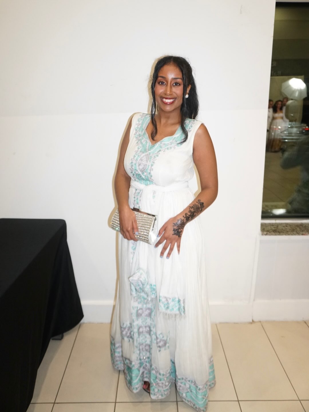 A woman wearing a sleeveless white maxi Habesha dress with light blue and green floral patterns, holding a metallic clutch in an indoor setting.