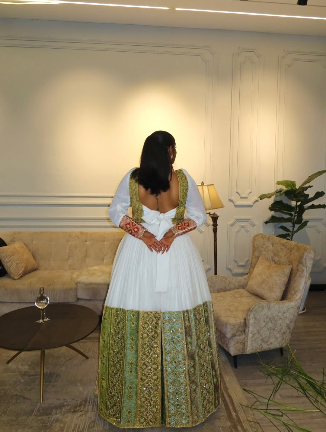 Woman in a white and green traditional Ethiopian dress standing in a stylish room with decor elements.