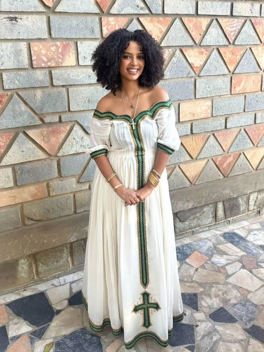 Woman in a white green Habesha Libs with green accents standing against a patterned wall.