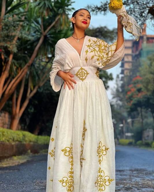 Woman in a white Ethiopian cultural dress dress with gold embroidery holding flowers outdoors.