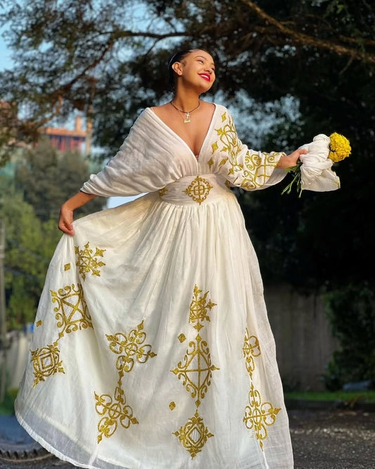 Woman in a white Ethiopian cultural dress with gold embroidery standing outdoors.
