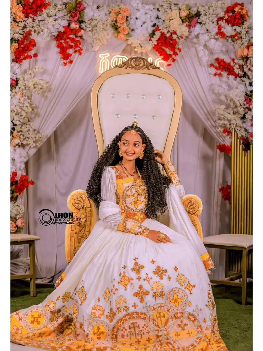 Woman in a white and gold embroidered Gold Ethiopian Wedding Dress sitting on an ornate chair with floral decorations in the background.