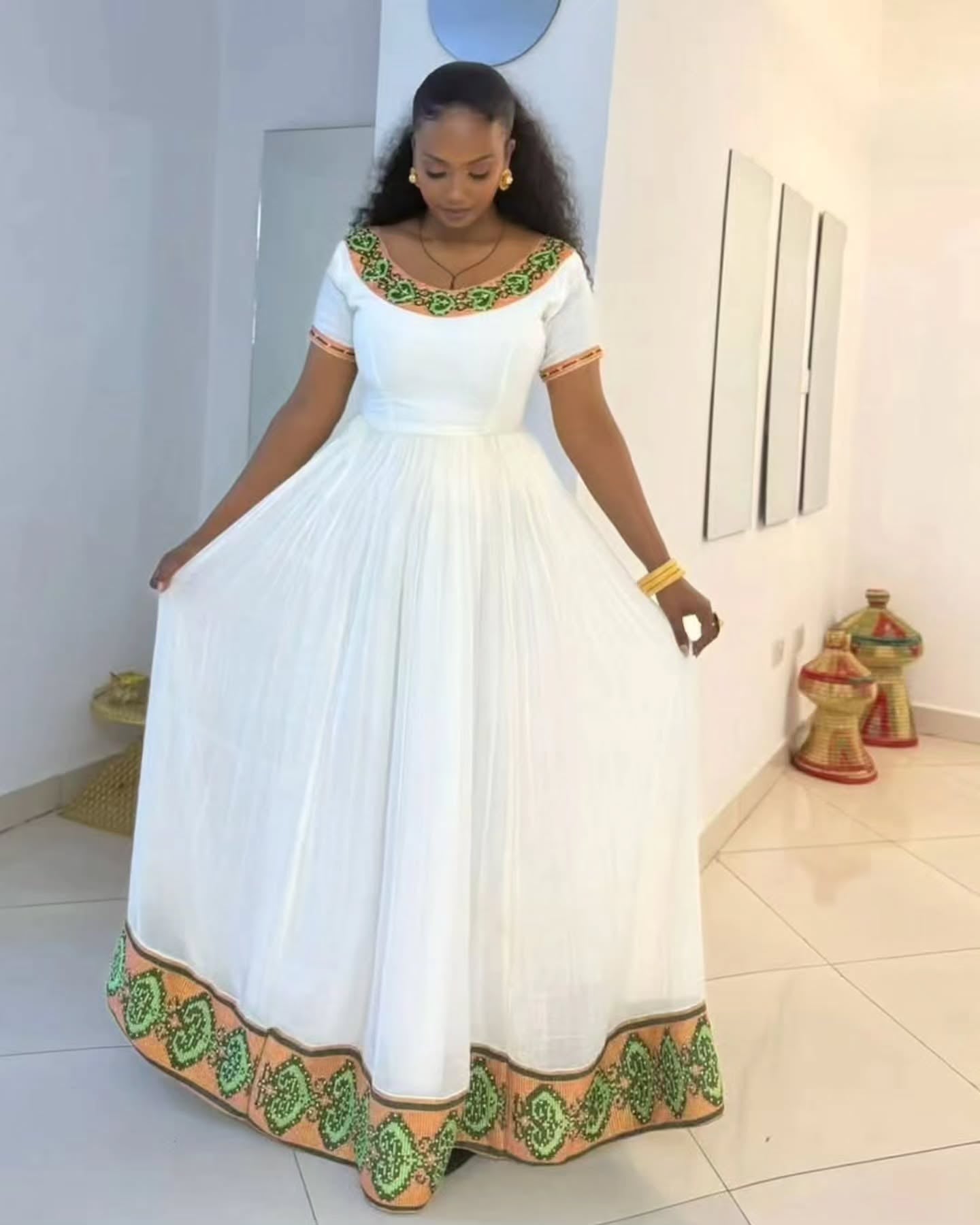 A woman modeling a white Habesha Kemis with green and orange embroidered patterns on the neckline, sleeve cuffs, and hem, standing in a brightly lit indoor room.