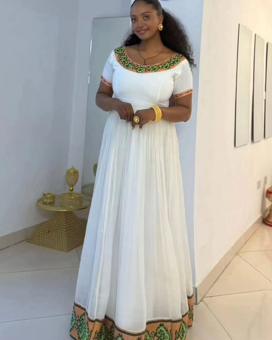 A woman wearing a white Habesha Kemis with green and orange embroidered details on the neckline and hem, standing in a brightly lit indoor hallway.