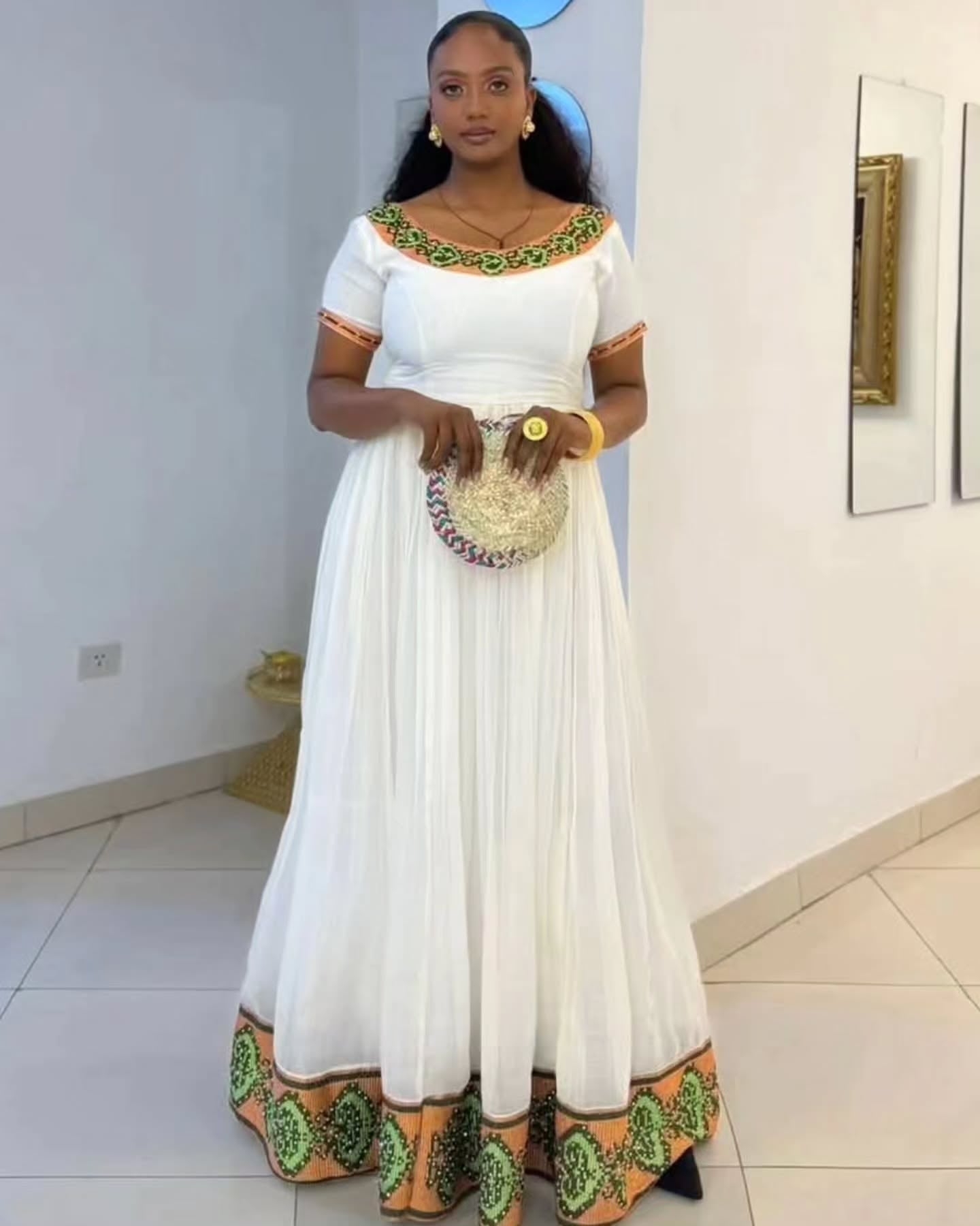 A woman modeling a white Habesha Kemis with green and orange printed border details on the neckline, sleeves, and hem, holding a woven clutch purse in an indoor setting.