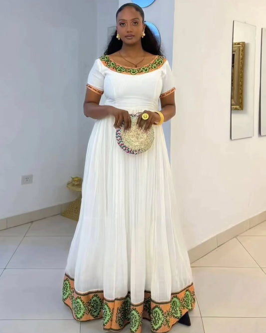 A woman modeling a white Habesha Kemis with green and orange printed border details on the neckline, sleeves, and hem, holding a woven clutch purse in an indoor setting.