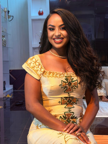 A woman with long, wavy dark brown hair is smiling and wearing a cream-colored traditional Ethiopian Traditional Clothing with gold embroidery and green accents. She is seated in an indoor setting with shelves and furniture visible in the background.