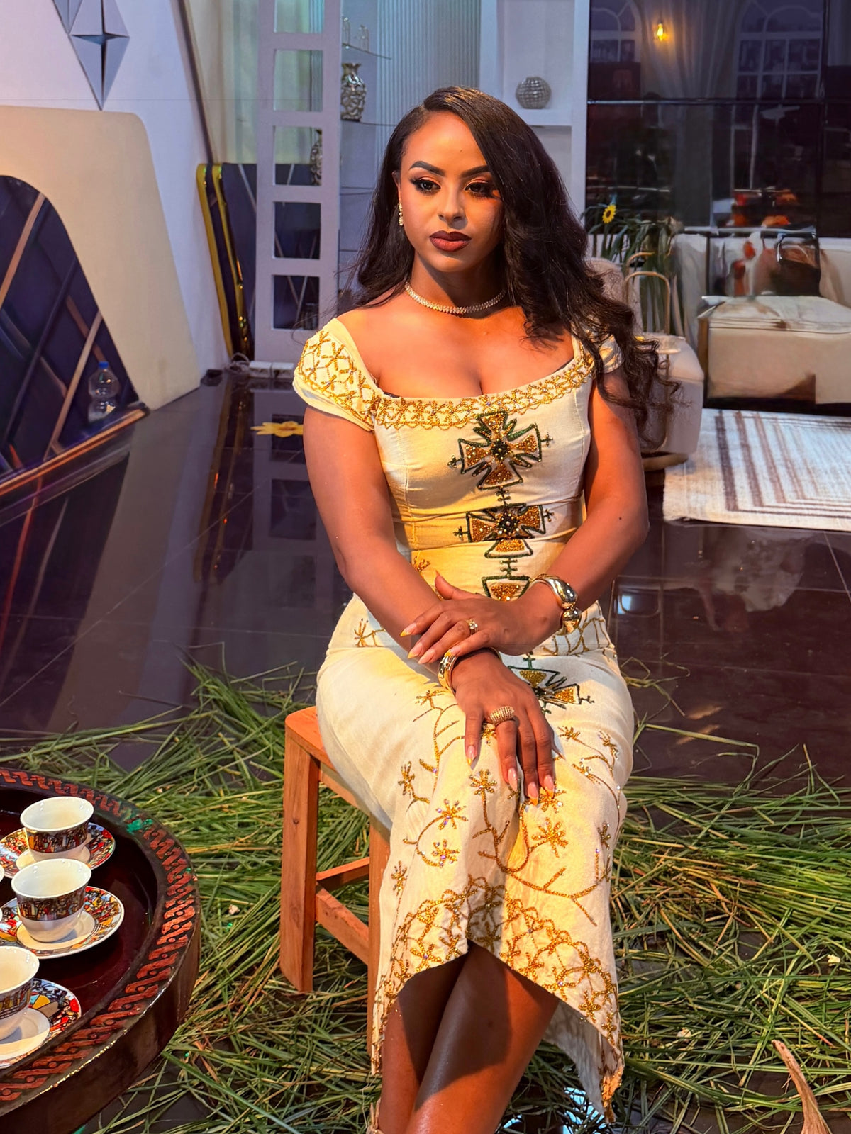 A woman sits on a wooden stool wearing a cream and gold embroidered Ethiopian Traditional Clothing in an indoor setting with a dark reflective floor and decorative greenery.