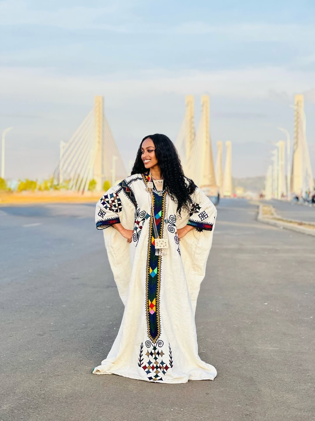 Woman in a white Gondar Fetel with colorful patterns standing on a road with modern architecture in the background