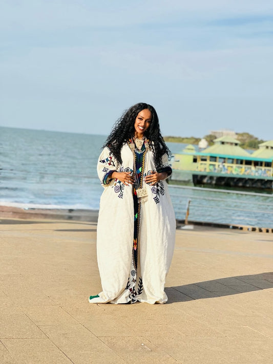 Woman in a long, white Gondar Fetel with decorative patterns standing by a waterfront.