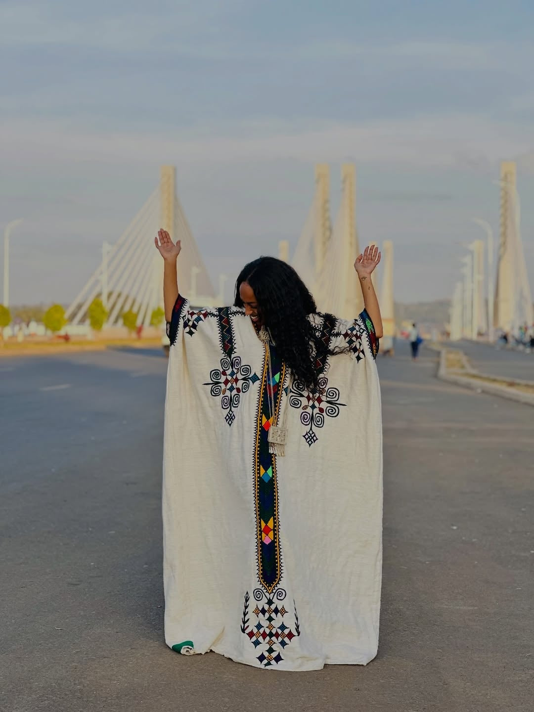 Woman in a white Gondar Fetel with black embroidery standing on a road with a bridge in the background