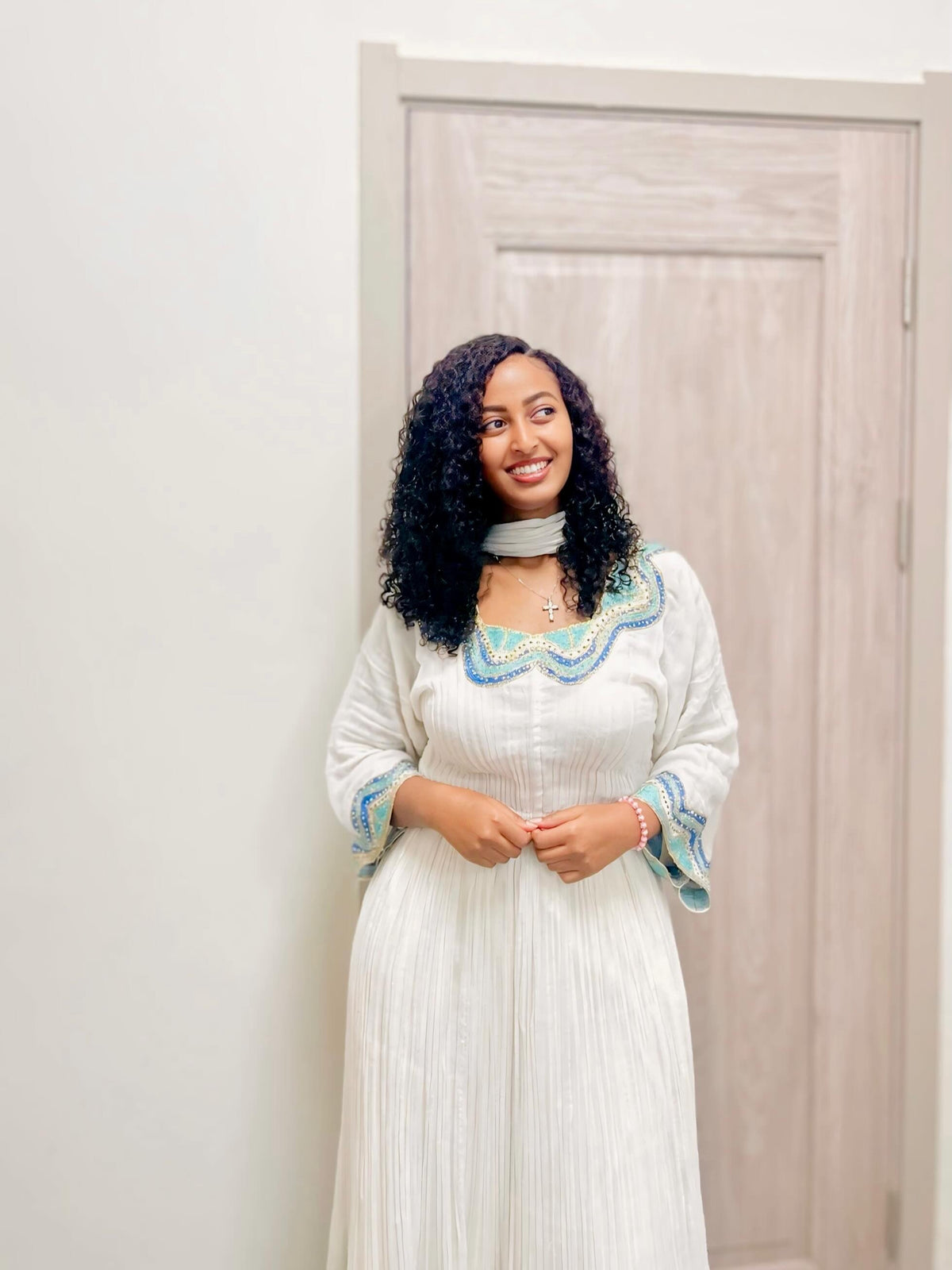 A woman with curly black hair stands indoors wearing a long white  Habesha kemis  with blue and green embroidered detailing around the neckline and cuffs, paired with a light gray choker necklace.
