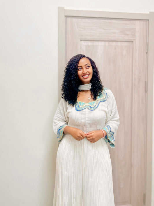A woman with curly black hair stands indoors wearing a long white  Habesha kemis  with blue and green embroidered detailing around the neckline and cuffs, paired with a light gray choker necklace.