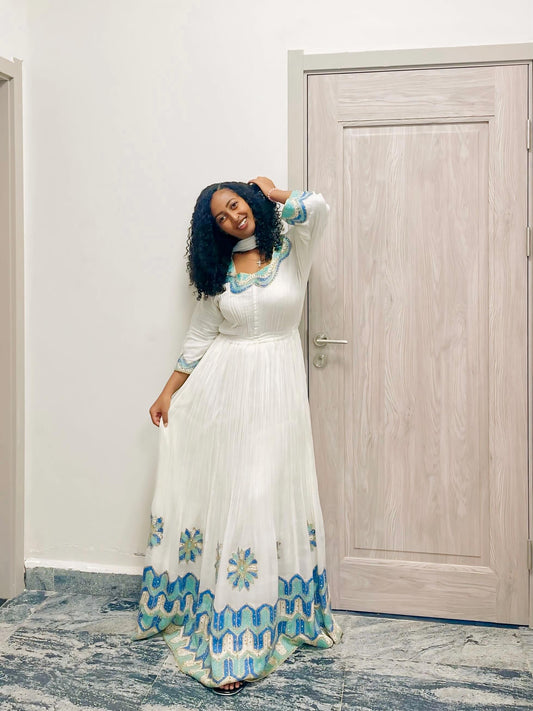 A woman modeling a white  Habesha kemis  with blue and green patterned accents at the neckline and hem, standing in an indoor setting next to a light wood door.