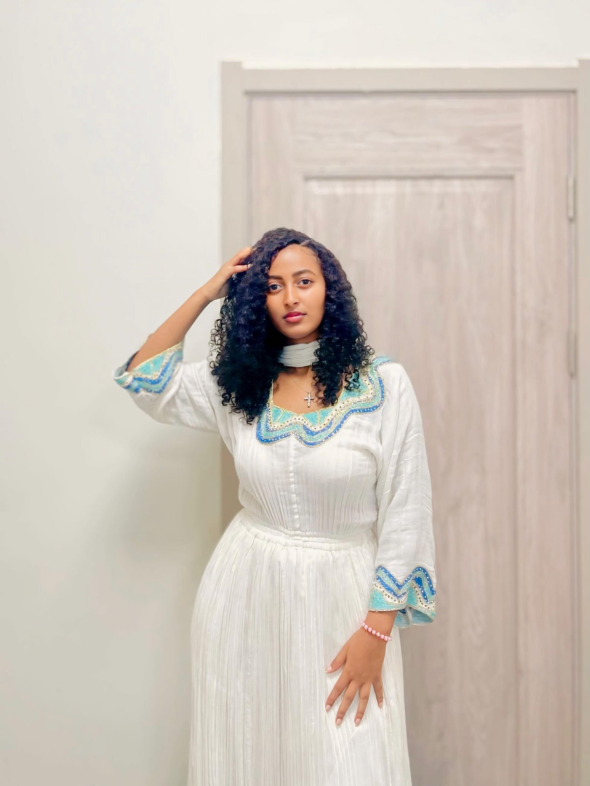 A woman with curly black hair models a white long-sleeve  Habesha kemis  with blue and green embroidered details on the collar and cuffs, standing in front of a light-colored wooden door.