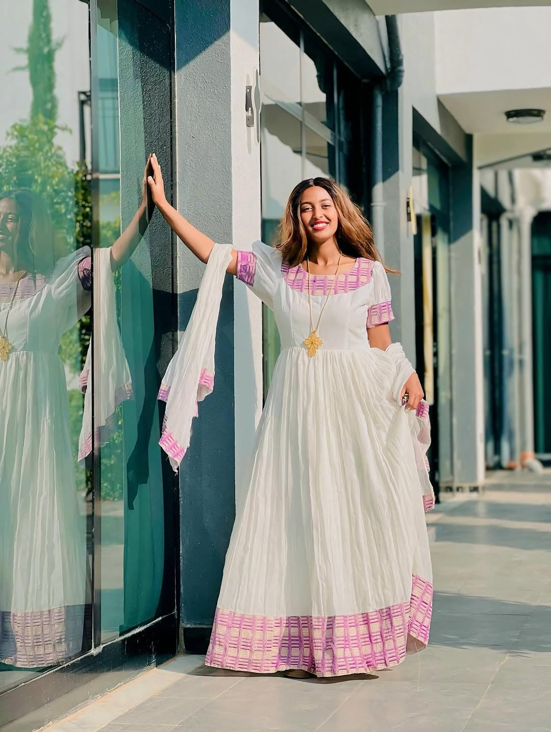 Woman in a white and pink traditional habesha kemis standing in front of a glass building.