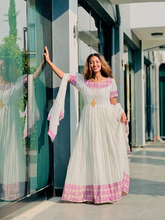 Woman in a white and pink traditional habesha kemis standing in front of a glass building.