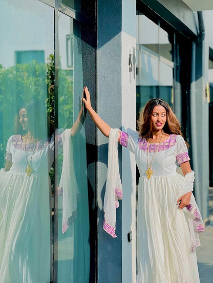 Woman in a white Habesha Kemis with pink accents standing next to a reflective surface.