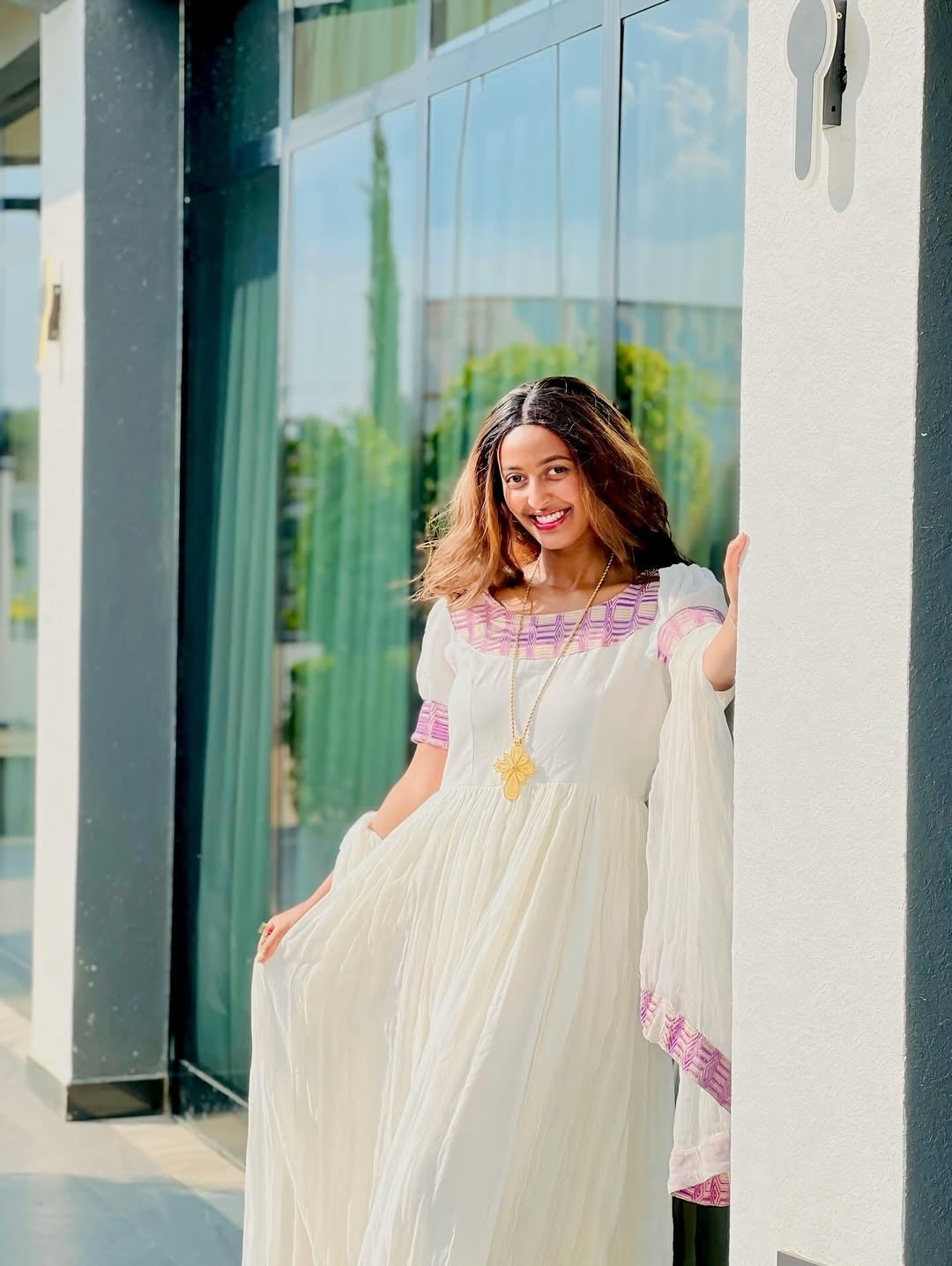 Woman in a white dress with pink blouse standing outdoors near a building.
