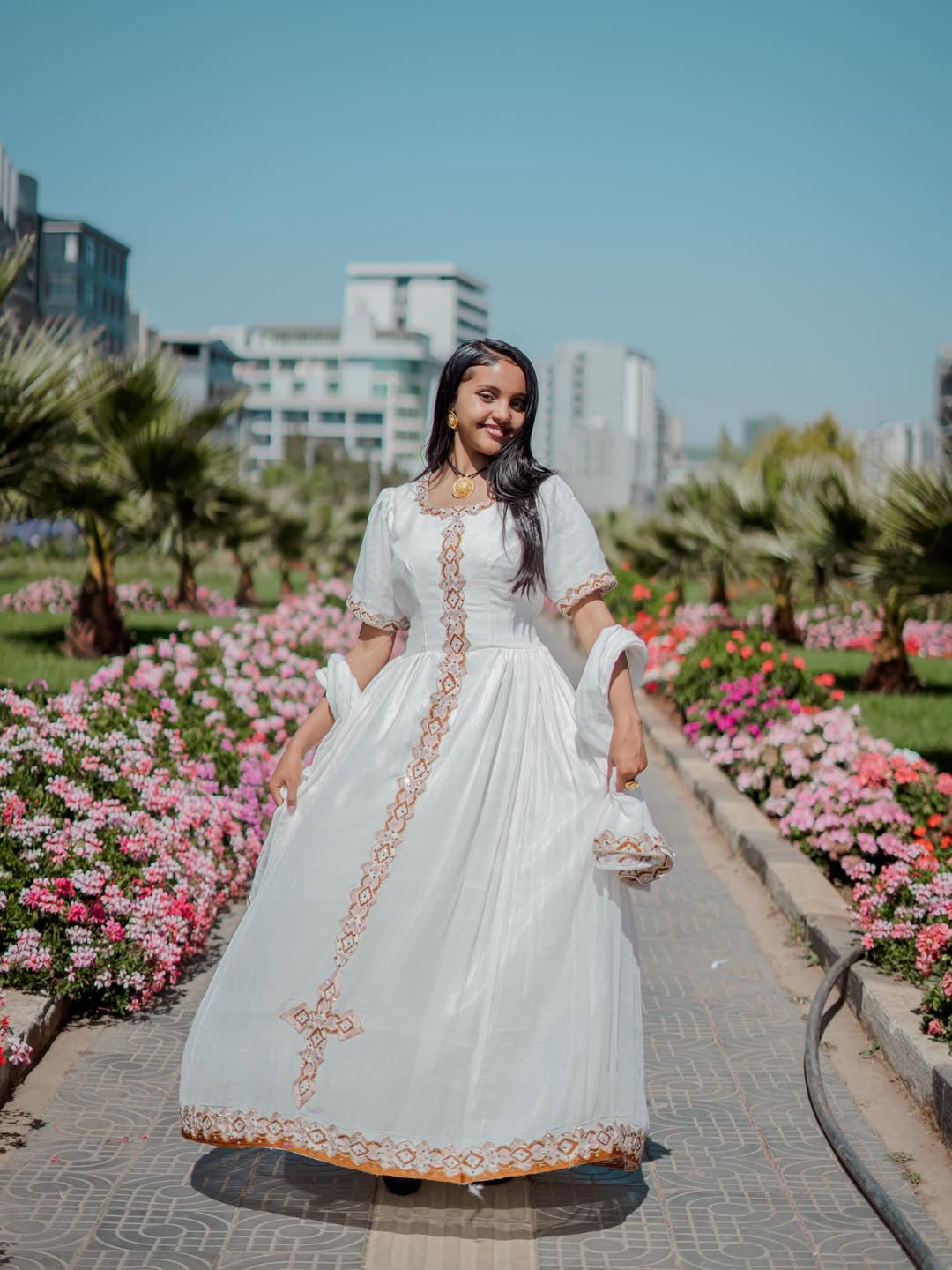 Woman in a white traditional habesha dress with gold embroidery standing in a garden with flowers and buildings in the background.