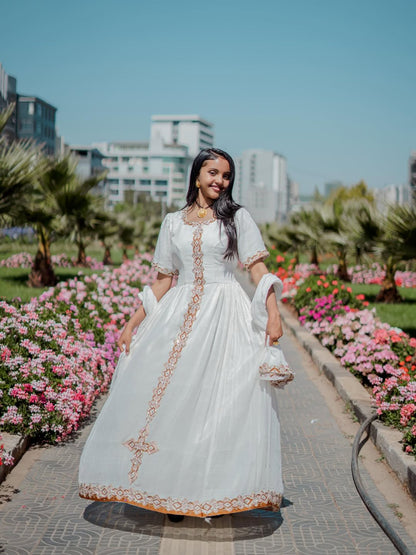 Woman in a white traditional habesha dress with gold embroidery standing in a garden with flowers and buildings in the background.