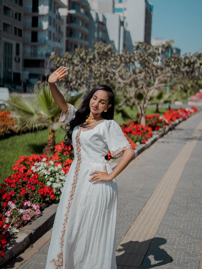 Woman in a habesha dress standing on a sidewalk with flowers and buildings in the background