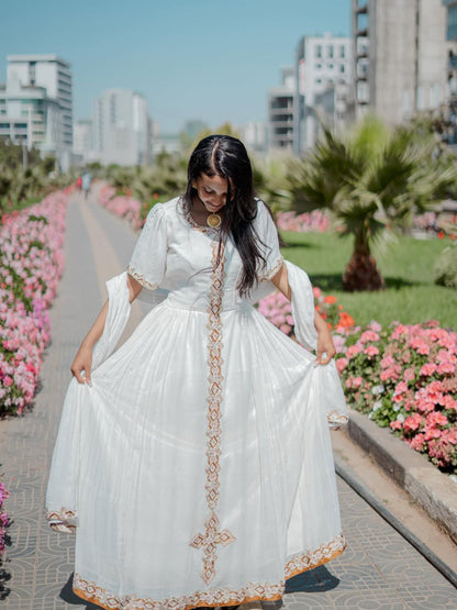 Woman in a white habesha dress with gold embroidery walking in a park with flowers and buildings in the background