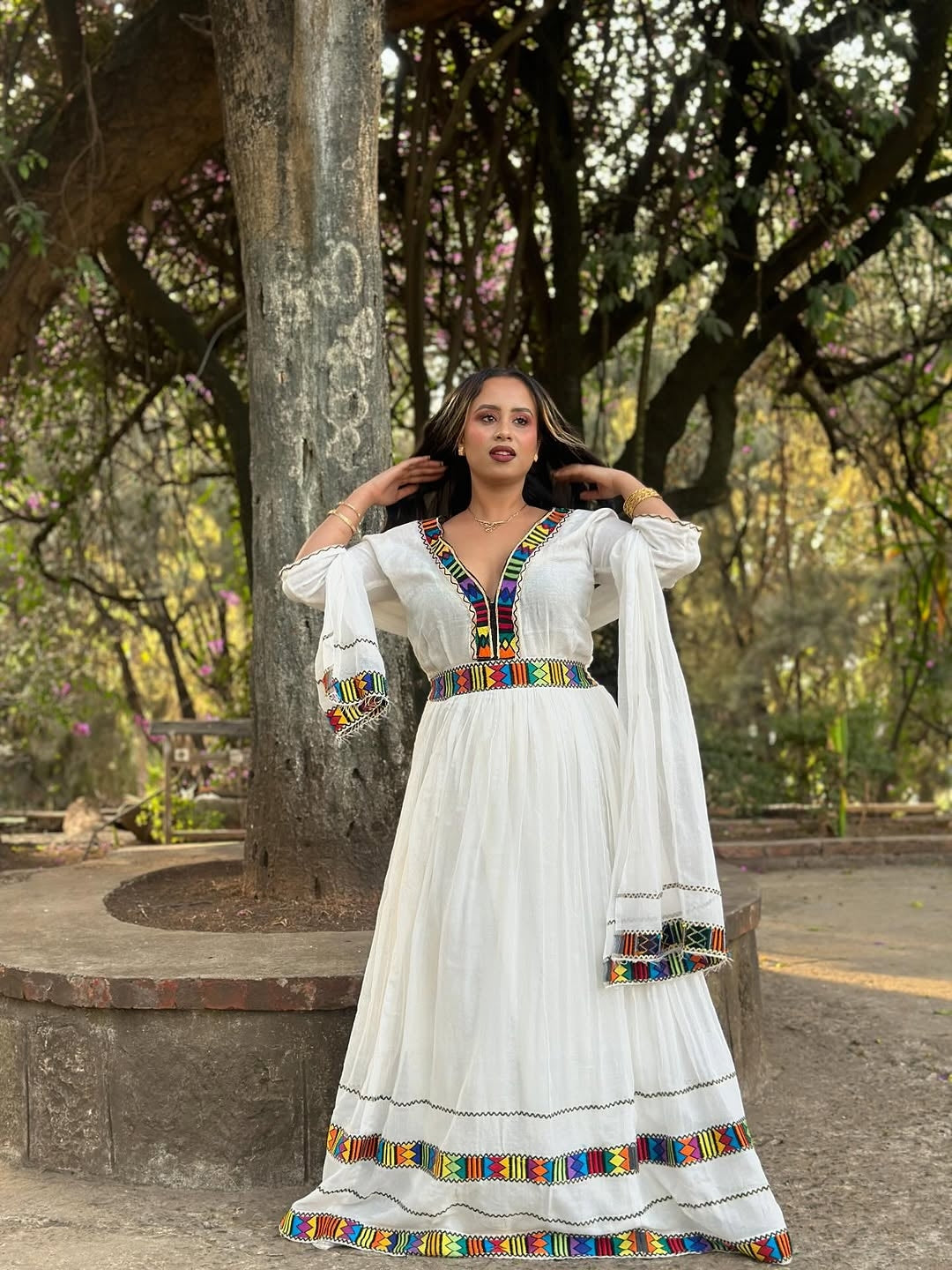 Woman in a white Habesha Kemis with colorful embroidery standing outdoors near trees.